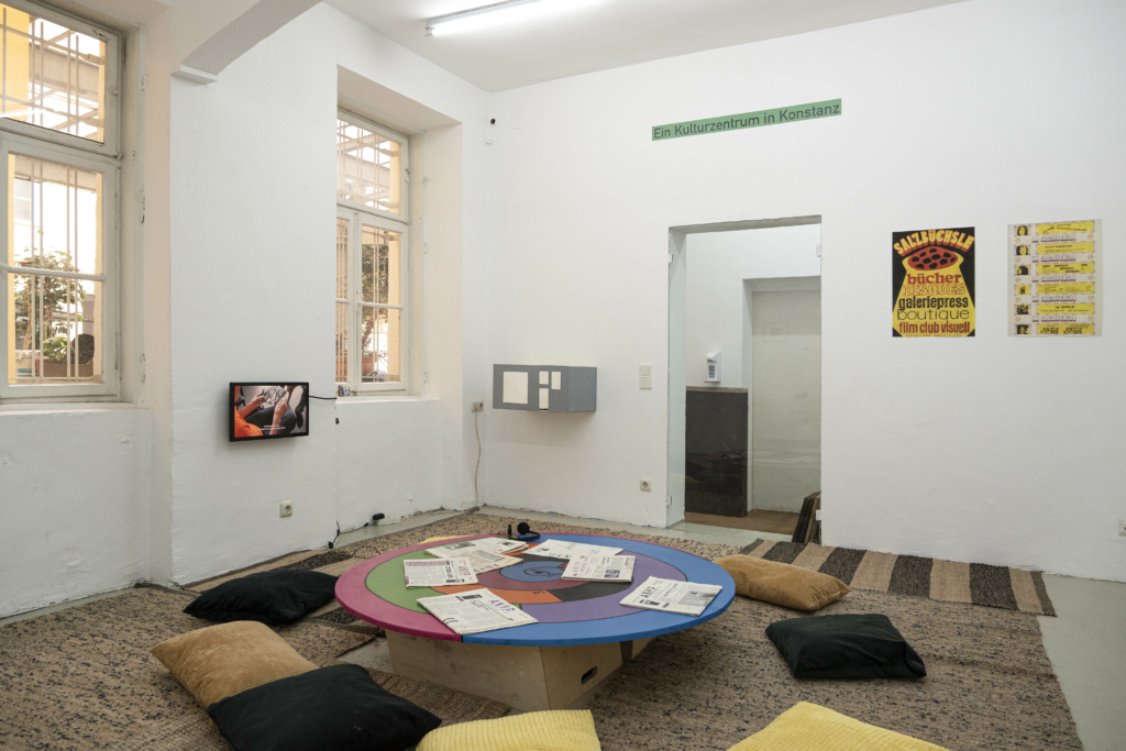 Raumansicht, in der Mitte der bunte spiralförmige Tisch mit Zeitungen, darum Kissen. An den Wänden ein Fernseher, eine graue Box und gelbliche Plakate. Über allem der Schriftzug "Ein Kulturzentrum in Konstanz".