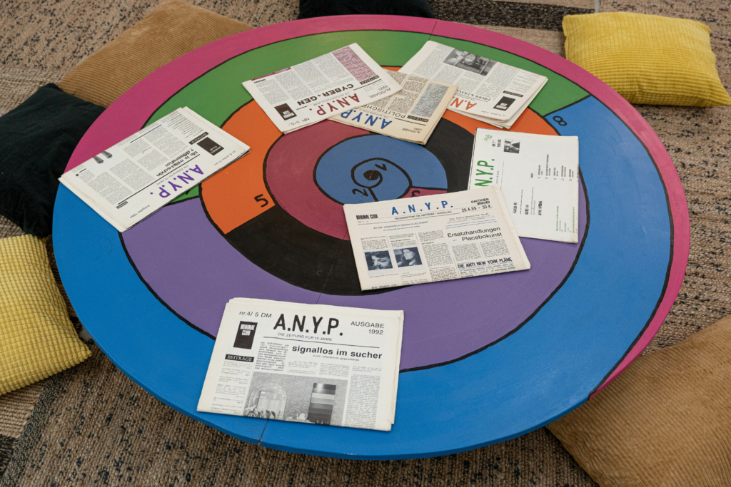 Ein runder, spiralförmig bunter Tisch knapp über dem Boden, auf welchem Zeitungen mit dem Namen "A.N.Y.P." liegen.