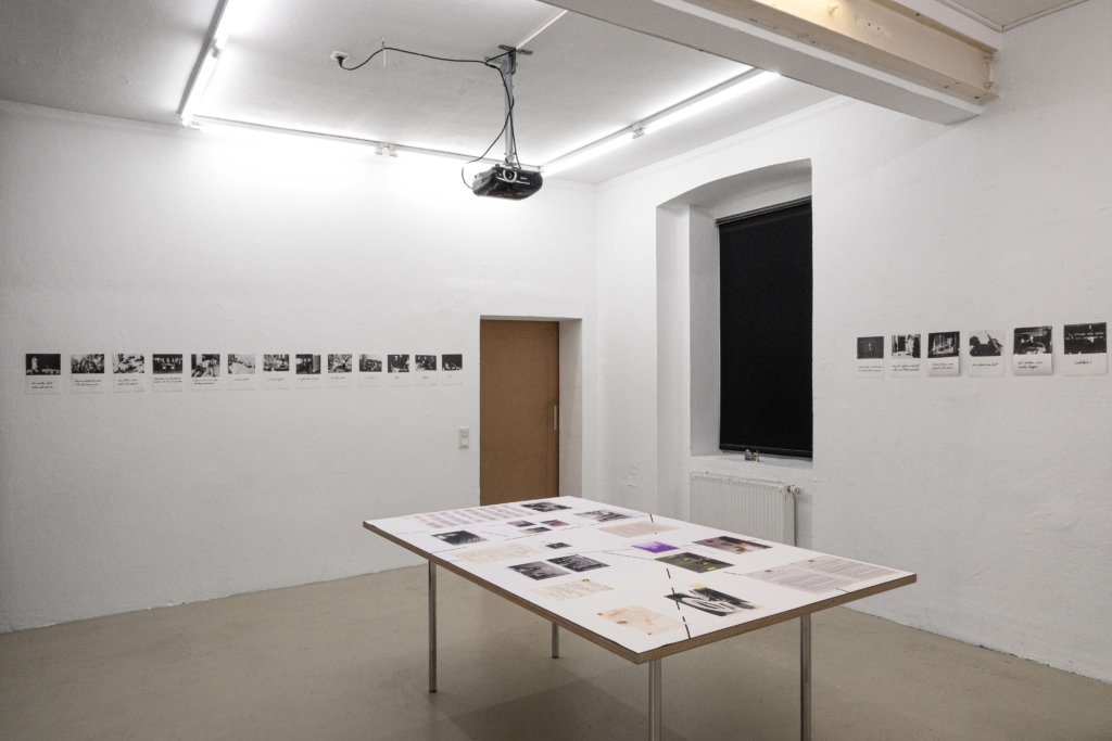 Ausstellungsansicht: Ein weißer Raum mit einem Tisch in der MItte, auf welchem Fotos und beschriebenes Papier liegt. An den Wänden mehr schwarz-weiß Fotografien in einer Reihe.