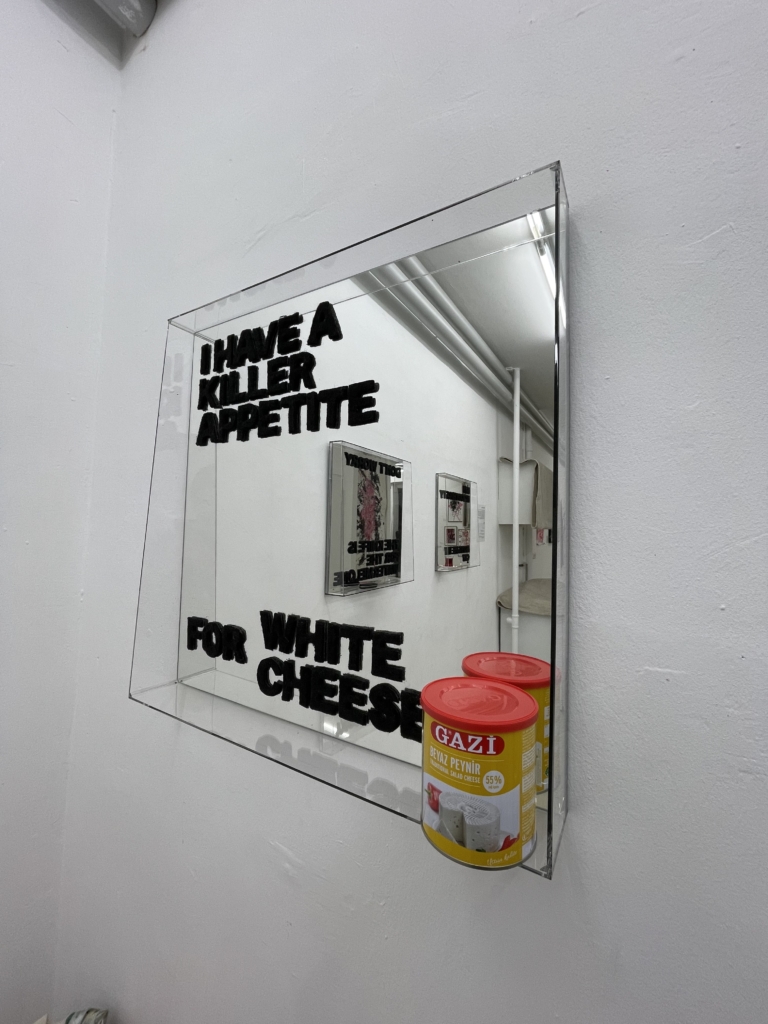 Ausstellungs-Nahansiccht eines Spiegels, der die Worte "I have a Killer Appetite for White Cheese" auf sich stehen hat. Unten rechts am Spiegel eine Dose Gazi Käse.