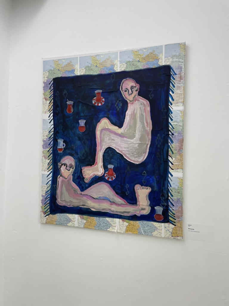 Foto eines Gemädes an der Wand einer Ausstellung: Zwei abstrakte geschlechtslose Figuren sitzen auf einer blauen Decke mit Chai-Tassen um sie verbreitet. Das Gemälde ist auf Landkarten von Deutschland gemalt.