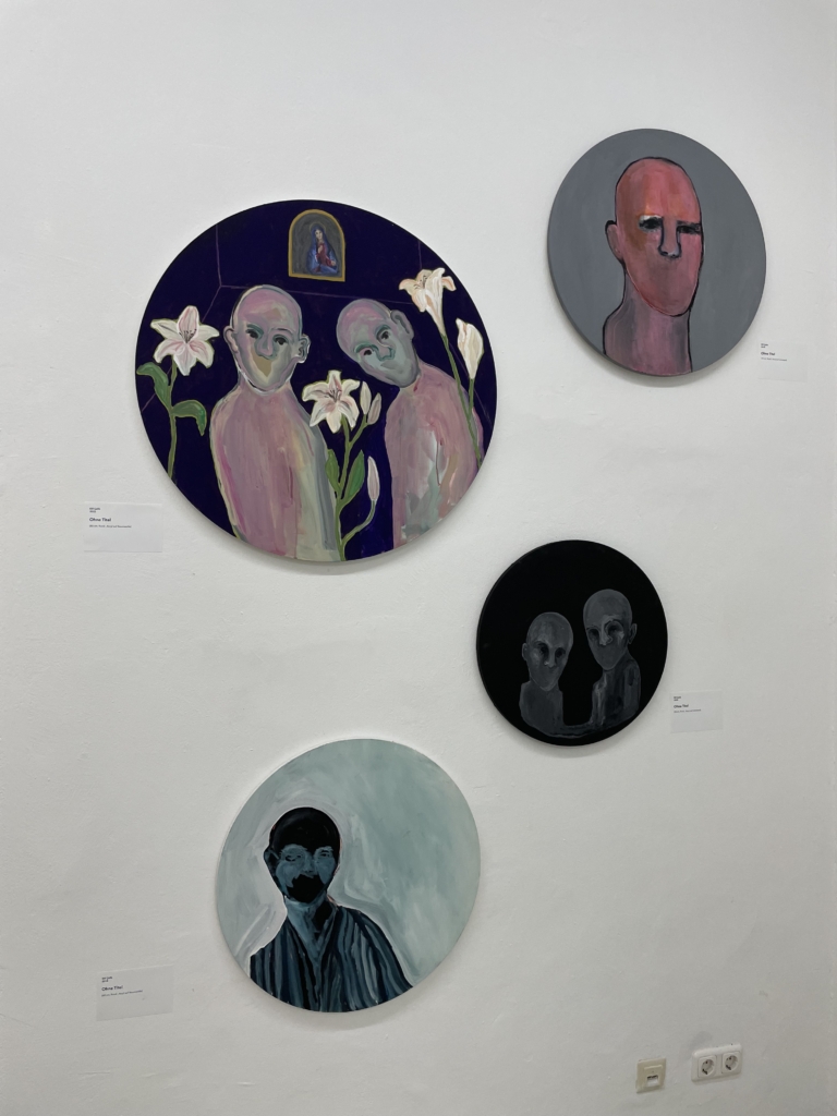 Nahaufnahme von vier runden Gemälden, die glatzköpfige, gecshlechtslose Figuren zeigen, die den Betrachtenden anschauen.