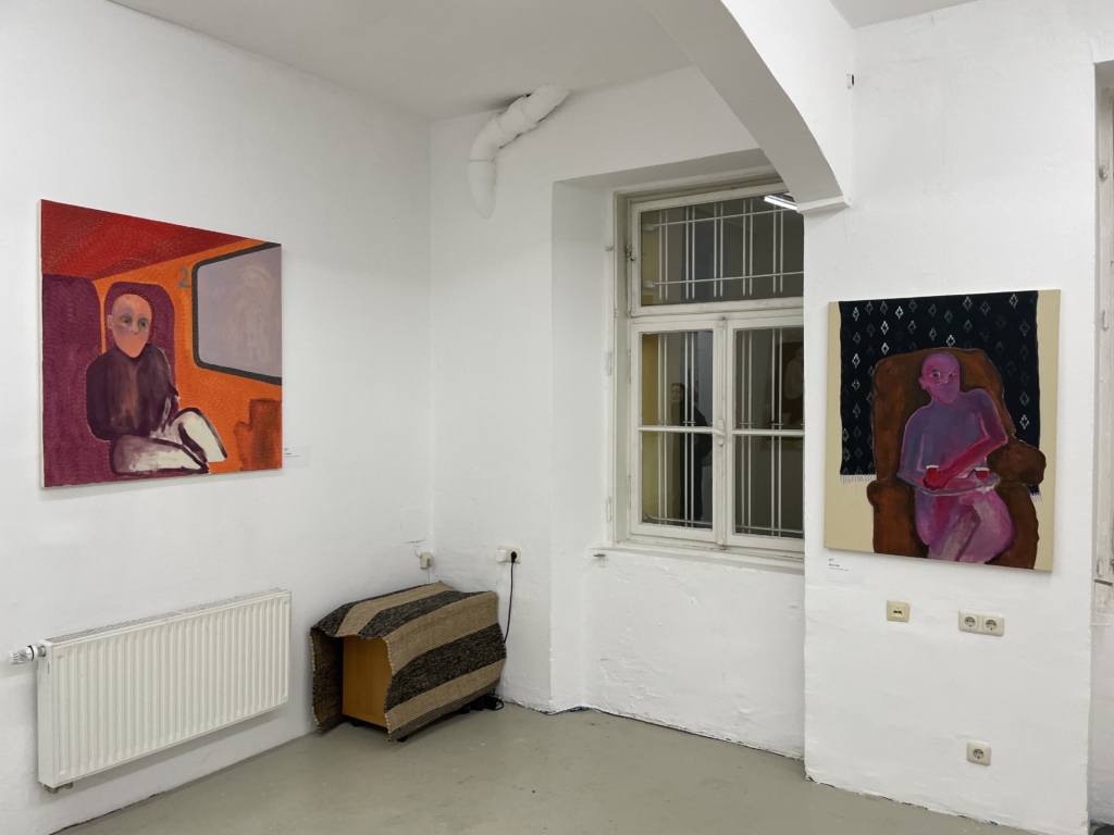 Ausstellungsansicht: Links das Gemälde in rot-orange mit einer abstrakten Person im Zugabteil. Rechts ein Gemälde einer abstrakten gecshlechtslosen Person auf einem brauenn Sessel mit einem Tablett mit zwei Chai-Gläsern. Dazwischen das Fsnter und eine Teppichbedeckte Truhe.