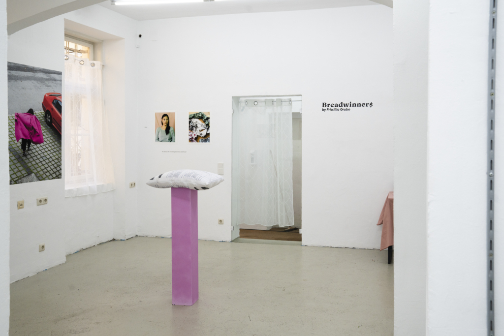 Ausstellungsansicht der Ausstellung "Breadwinners" von Priscillia Grubo" In der Mitte des Raumes ist ein pinker Sockel mit einem Kissen darauf, an den Wänden Fotografien verschiedener Größen, die Frauen zeigen. Eine Tür führt in einen anderen Raum.