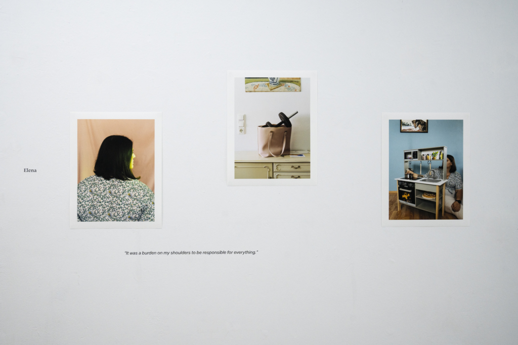 Direkt-Aufsicht auf Wand in Ausstellung. Drei Fotografien zeigen eine Frau von hinten, eine Handtasche auf einer Kommode mit einer Hantel darin und eine Kinderspielküche mit einer Frau dahinter. Links an der Wand steht der Name "Elena".