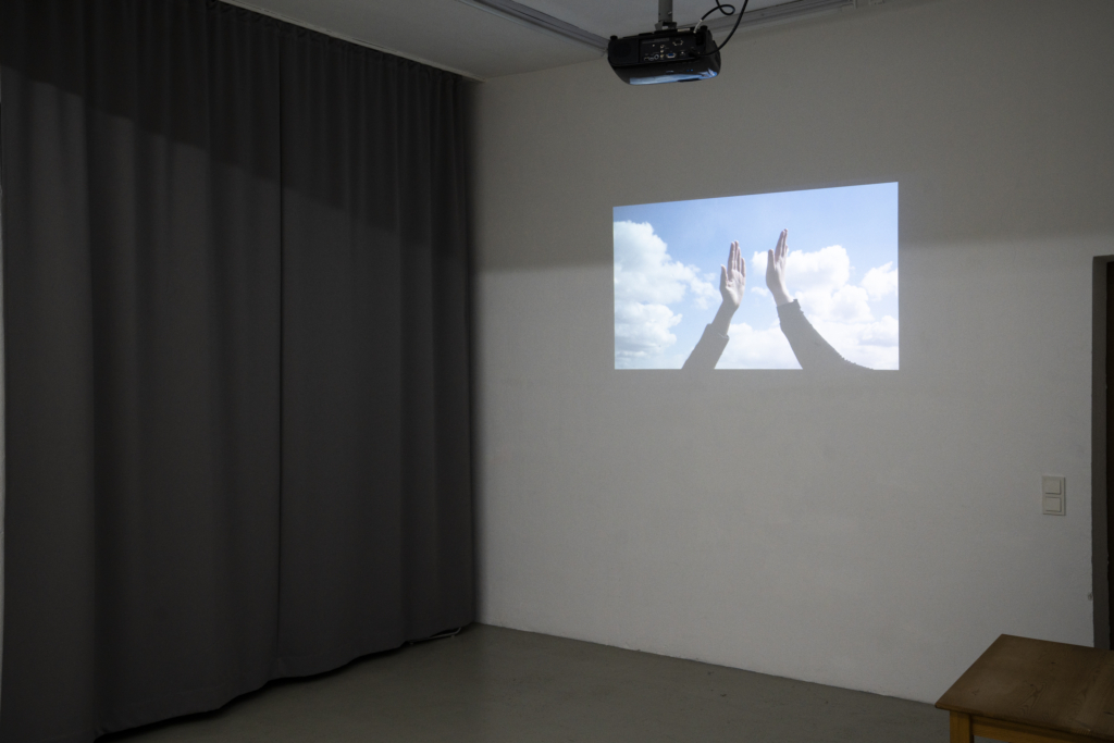 Foto: Ausstellungsansicht einer Projektion an der Wand, die zwei klatschende Hände vor dem wolkigen Himmel zeigt. Links neben der Projektion ein grauer Vorhang.