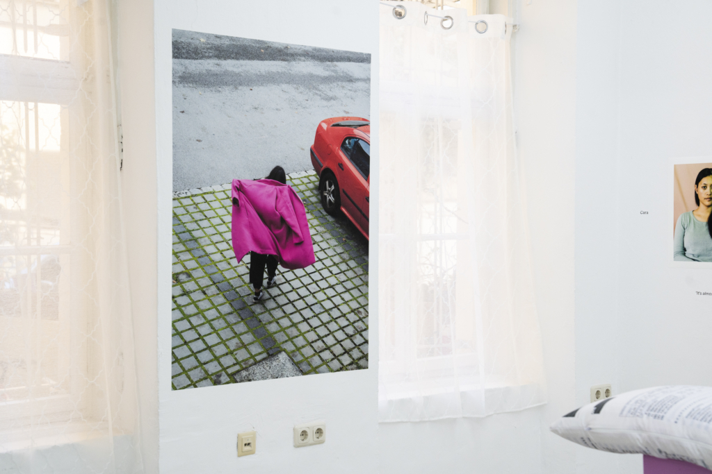 Foto: Ein großer Foto-Print einer Frau, die einen knalligen pinken Mantel anzieht und auf einem Parkplatz an einem roten Auto vorbeigeht. Neben dem Print weiße Vorhänge, rechts angeschnitten ein vom Bild abgeschnittener Print einer Frau.