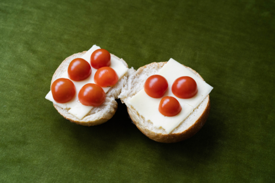 Das Bild zeigt ein aufgeschnittenes helles Brötchen auf einem dunklen Hintergrund. Beide Brötchenhälften sind belegt mit einer Scheibe Käse und halbierten Tomaten.