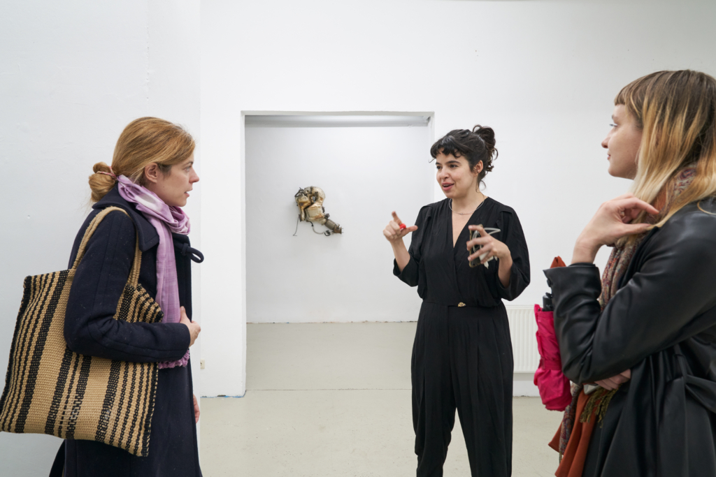 Drei junge Frauen im Gespräch, im Hintergrund ein Türbogen und eine Skulptur, die an der Wand hängt.
