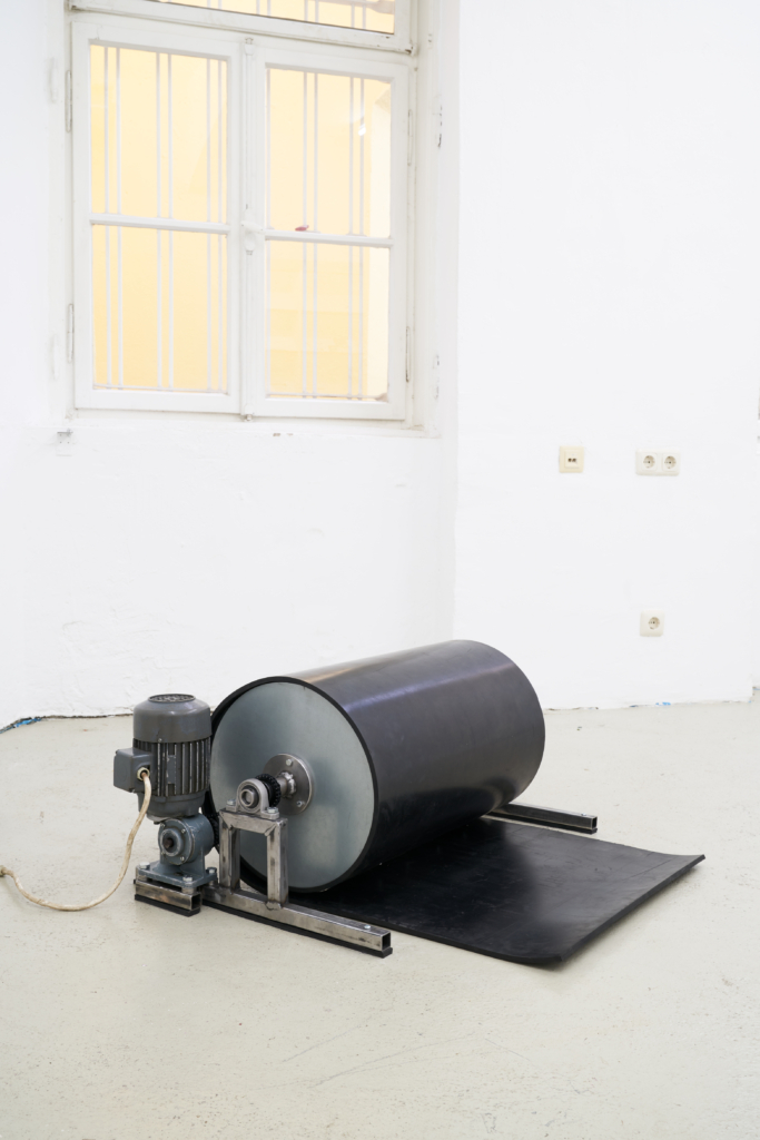 Foto: Weißer Galerieraum, auf dem Boden eine walzenartige Maschine mit Motor. Darüber ein Fenster.