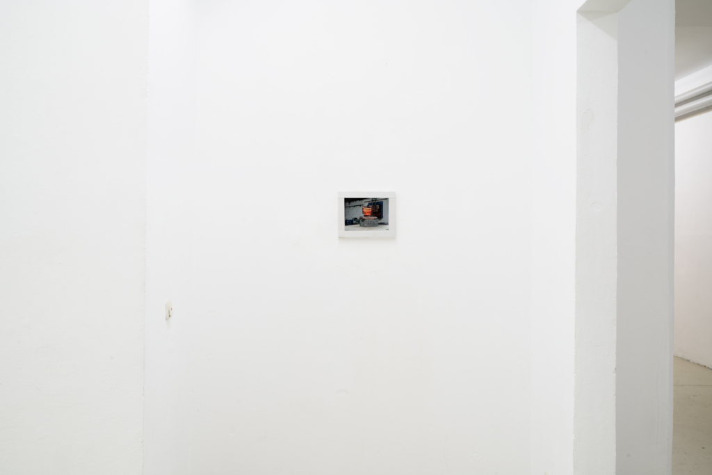 Foto: Weiße Galeriewand mit einem winzigen weiß gerahmten Bild, das etwas kleines rotes zeigt. Nicht genauer erkennbar.