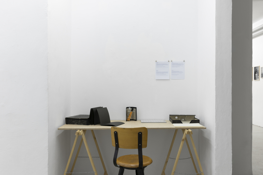 Foto einer Raumecke mit Holzböcken und Holztischplatte, darauf Schreibtischutensilien wie ein Aktenordner, Stifte, Papier etc. Davor ein Holzstuhl, an der Wand zwei beschriebene Papiere.