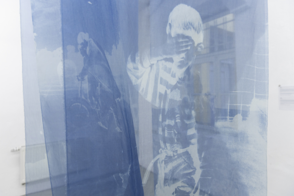 Nahaufnahme einer durchsichtigen Gaze, in weiß udn blau, die von der Decke hängt und das Portrait eines sich das Gesicht verdeckenden JUngen zeigt.