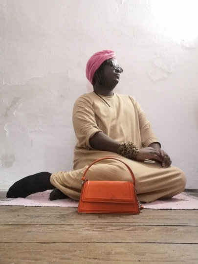 Photo of Keith Zenga King in the FLORIDA Space. Keith ists in a pose infront of a wall with an orange purse infront of him, wearing a beige dress and a pink cloth around the head plus black sunglasses.