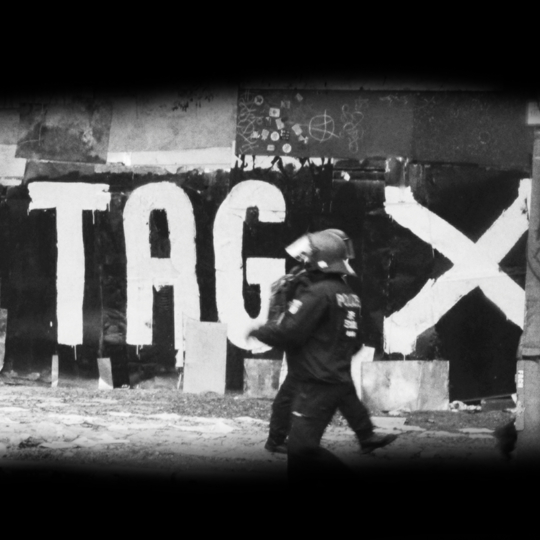 Zu sehen ist eine Schwarz-Weiß Fotografie aus dem Film. Zwei Polizisten stehen auf einer Straße in voller Einsatzmontur. Sie stehen vor den Toren des Köpi Wagenplatzes in Berlin. An der Wand ist mit Farbe in groß der Text Tag X hingemalt. Das Bild ist entstanden im Kontext der Räumung des Areals.